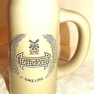 Puede incluir: Una jarra de cerveza de cer&aacute;mica beige con un logotipo de Heineken marr&oacute;n y un gr&aacute;fico de molino de viento. El logotipo dice "Heineken" y "Desde 1592".