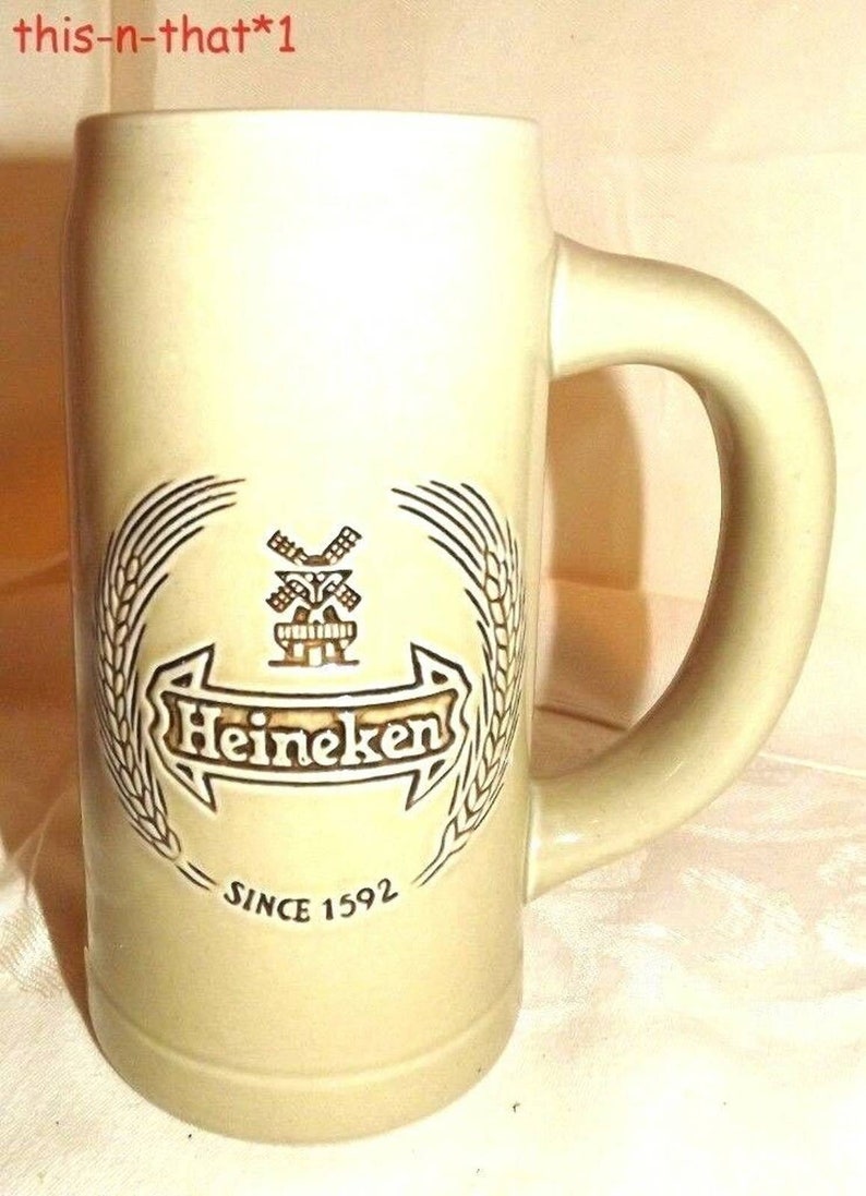 Puede incluir: Una jarra de cerveza de cer&aacute;mica beige con un logotipo de Heineken marr&oacute;n y un gr&aacute;fico de molino de viento. El logotipo dice "Heineken" y "Desde 1592".