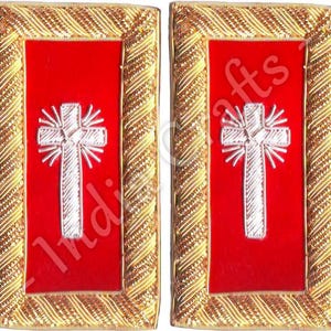 Puede incluir: Dos parches rectangulares con un centro rojo y un diseño de cruz blanca. La cruz está bordada con hilo plateado y rodeada por un borde dorado con un patrón tejido.