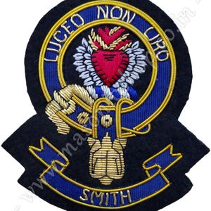 May include: A black felt embroidered patch with a gold and blue border. The center features a gold heart with a red flame and a gold crown. The text "LUCEO NON URO" is written in gold around the heart. The bottom of the patch has a gold ribbon with the text "SMITH" in gold.