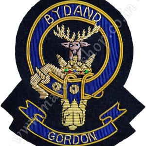 May include: A black felt embroidered patch with gold thread depicting a stag's head with a crown and the text "BY AND GORDON".