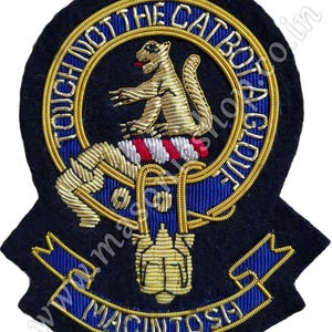 Pode incluir: Um remendo bordado azul e dourado com o texto "Touch Not The Cat Bot Ago" e "MacIntosh" em um círculo. O remendo apresenta um animal bordado em ouro e prata.