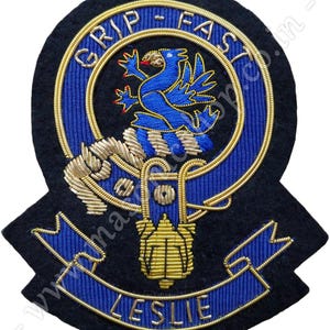 Puede incluir: Parche bordado negro con un emblema de pájaro azul y las palabras "GRIP - FAST" en letras doradas. El parche también presenta el nombre "LESLIE" en una pancarta azul, con detalles dorados y azules.