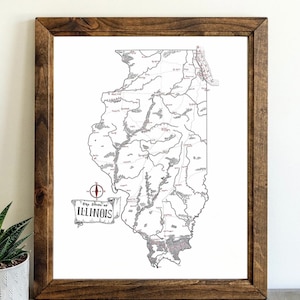 May include: A framed map of Illinois, featuring a detailed black and white illustration of the state's geography. The map includes rivers, towns, and a compass rose. The title "The State of Illinois" is displayed on a banner. The frame is made of dark brown wood.