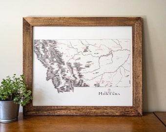 May include: A framed map of the state of Montana with the names of towns and cities printed in black text. The map is on a white background and has a brown wooden frame.