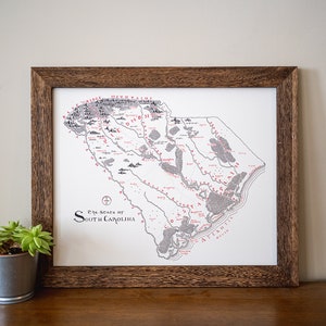May include: A framed black and white map of the state of South Carolina with red text. The map shows rivers, towns, and other geographical features. The text at the bottom reads "The State of South Carolina".