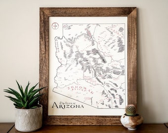 May include: A framed map of Arizona with the text "The State of Arizona" and "Sonoran Desert" in red lettering. The map is filled with black and white text and lines. The frame is brown wood.