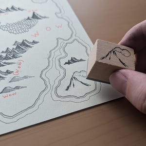 May include: A wooden stamp with a black and white illustration of a mountain range with a cloud above it. The stamp is being held by a hand over a map with a similar illustration.