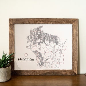 May include: A framed map of the state of Wisconsin with the title "The State of Wisconsin" printed in the bottom left corner. The map is in black and white with red markings. The frame is made of dark wood.
