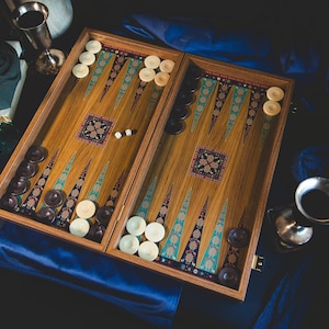 May include: A wooden backgammon set with a detailed inlaid design. The game board features brown and white checkers, dice, and decorative patterns in teal, red, and gold. The set is displayed on a dark blue surface, suggesting a game night theme.