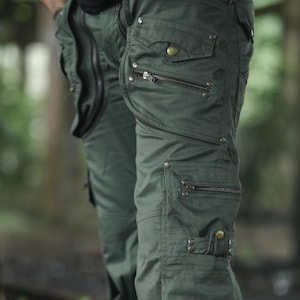 Op de afbeelding: Donkergroene cargobroek met meerdere zakken en ritsen. De broek heeft een militair geïnspireerd ontwerp, met verschillende compartimenten met ritsen en decoratieve metalen knopen. De stof lijkt een duurzaam, mogelijk waterafstotend materiaal te zijn.