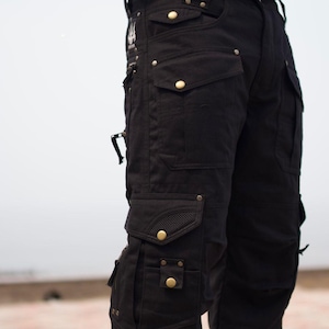 May include: Black cargo pants with multiple pockets and brass-colored button details. The pants have a utilitarian design with a rugged aesthetic, suitable for outdoor activities or casual wear.