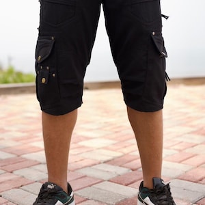 May include: Black cargo shorts with multiple pockets, zippers, and decorative rivets. The shorts are knee-length and worn with black, white, and green sneakers. The image is taken outdoors on a brick surface.