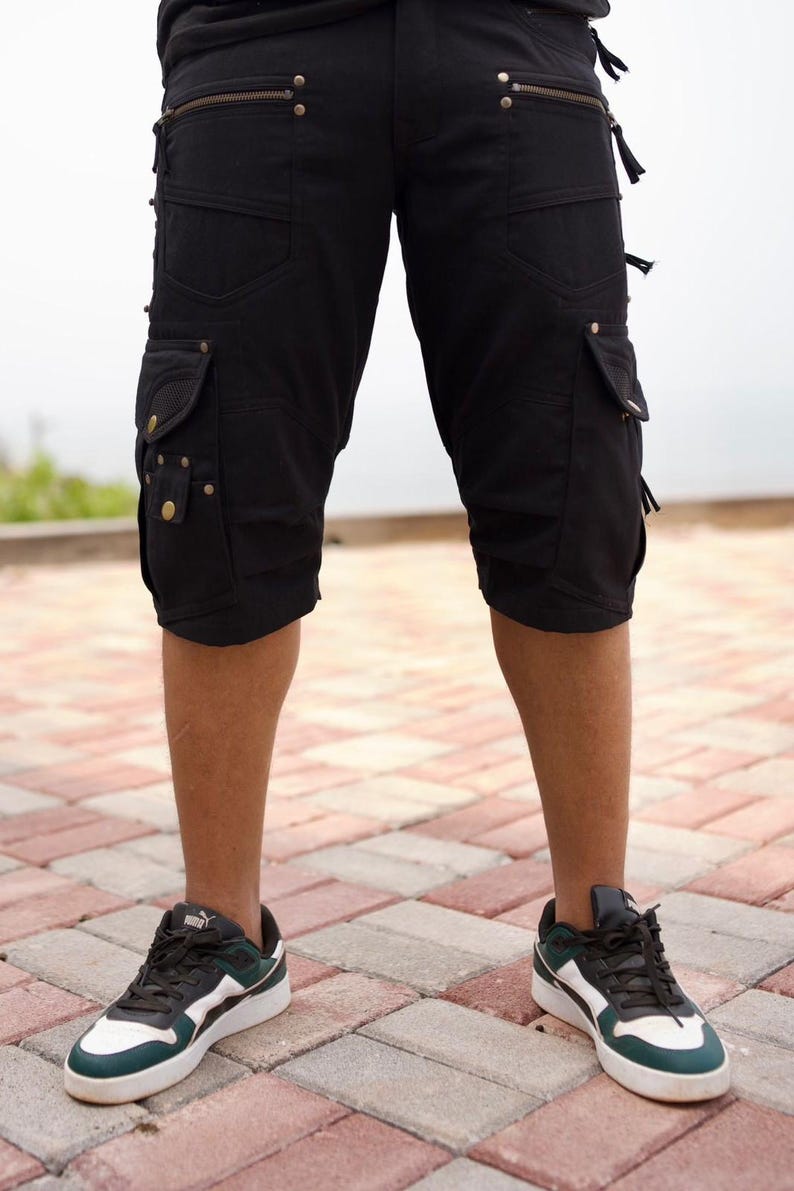 May include: Black cargo shorts with multiple pockets, zippers, and decorative rivets. The shorts are knee-length and worn with black, white, and green sneakers. The image is taken outdoors on a brick surface.