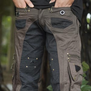 Op de afbeelding: Bruine cargobroek met meerdere zakken, ritsen en decoratieve metalen studs. De broek heeft een uniek ontwerp met een contrasterend donkergrijs stoffen paneel aan de voorkant van de benen. Het model draagt een zwart shirt.
