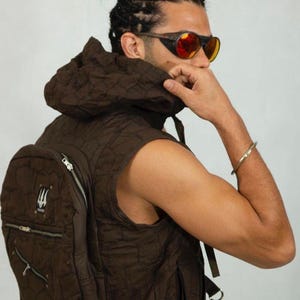 May include: A brown quilted vest with a matching backpack. The vest has a hood and a zipper closure. The backpack has multiple pockets and a logo. The person is wearing orange-tinted sunglasses and a gold bracelet.