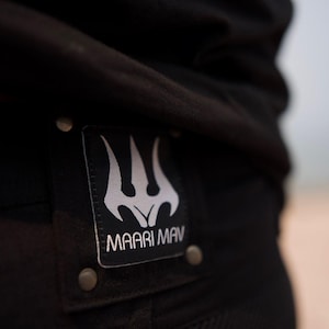 May include: Close-up of black clothing with a rectangular patch. The patch features a white trident-like design above the text "MAARI MAV". The patch is secured with metal rivets. The clothing appears to be made of a durable, textured fabric.