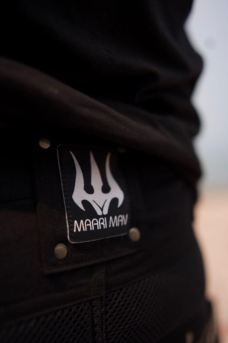 May include: Close-up of black clothing with a rectangular patch. The patch features a white trident-like design above the text "MAARI MAV". The patch is secured with metal rivets. The clothing appears to be made of a durable, textured fabric.