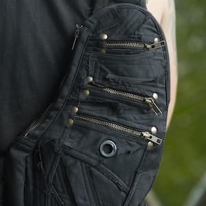 May include: Black shoulder bag with multiple zippered pockets and distressed fabric details. The bag features brass-colored rivets and zippers, and a circular grommet. The bag is worn over the shoulder.