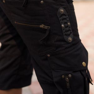 May include: Black cargo shorts with multiple pockets and zippers. The shorts feature metal rivets and a decorative leather accent. The design has a utilitarian aesthetic.