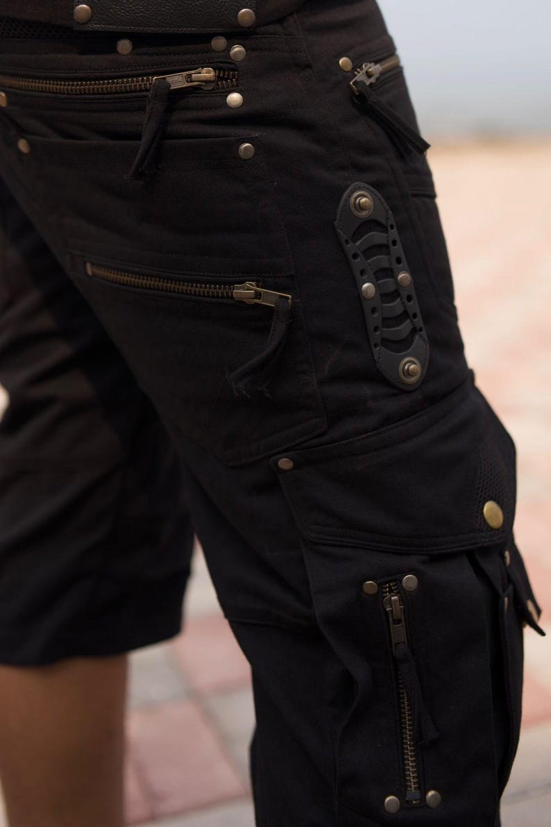 May include: Black cargo shorts with multiple pockets and zippers. The shorts feature metal rivets and a decorative leather accent. The design has a utilitarian aesthetic.