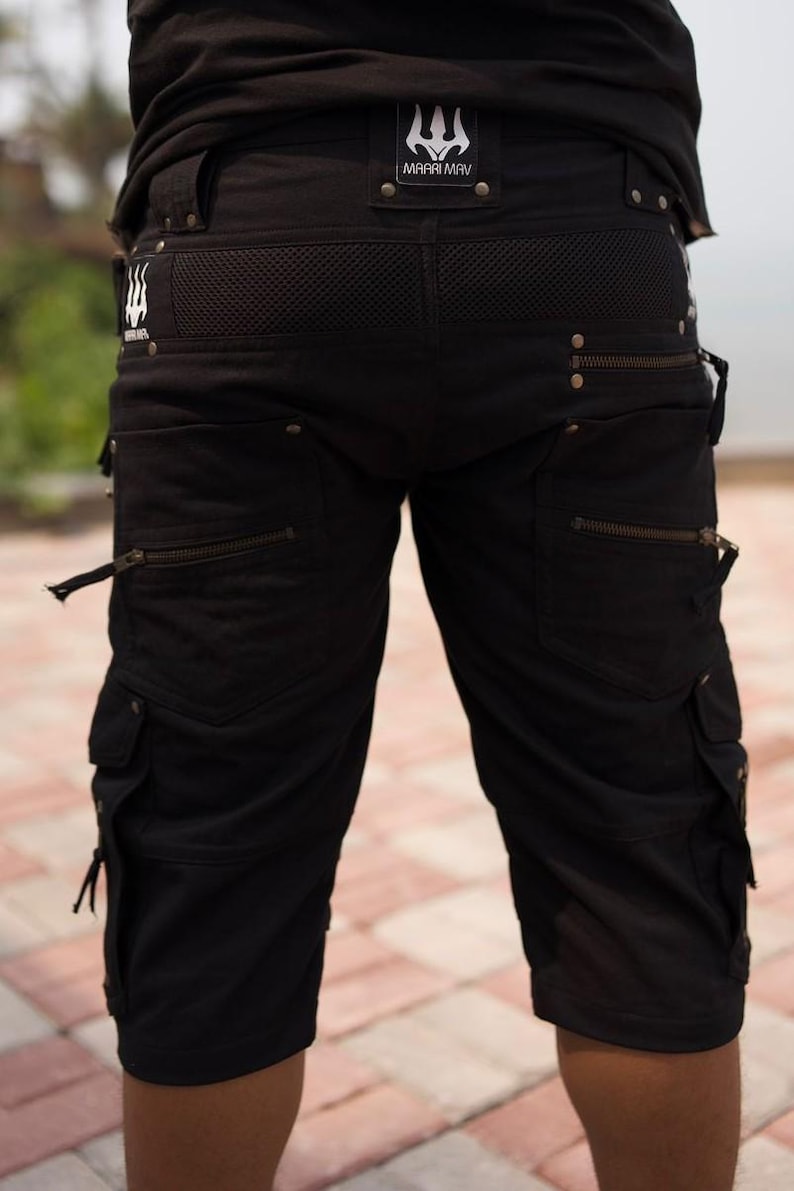 May include: Black cargo shorts with multiple pockets and zippers. The shorts feature a mesh panel at the back and a logo patch that reads "MAARI MAV". The shorts have a casual, yet rugged design.