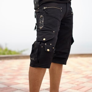 Puede incluir: Pantalones cortos cargo negros con múltiples bolsillos, cremalleras y remaches decorativos. Estos pantalones cortos hasta la rodilla tienen un diseño utilitario, adecuados para actividades al aire libre o uso casual.