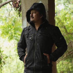 May include: A black zip-up hoodie featuring a subtle, intricate tonal pattern across the fabric. The garment includes a hood with drawstrings, multiple zippered pockets on the chest, and metallic stud accents along the seams and pocket edges.