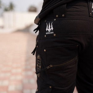 May include: Black cargo pants with multiple pockets, zippers, and metal rivets. The pants feature a white trident logo with the text "MAARI MAV". The pants are made of a durable fabric.
