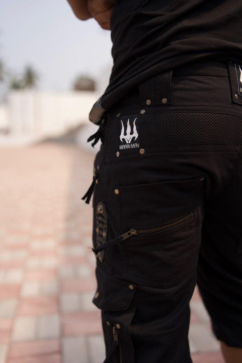 May include: Black cargo pants with multiple pockets, zippers, and metal rivets. The pants feature a white trident logo with the text "MAARI MAV". The pants are made of a durable fabric.