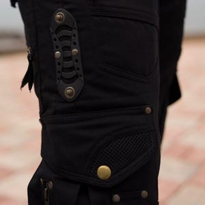May include: Close-up of black cargo pants with multiple pockets, zippers, and decorative metal rivets. The pants feature a unique design with a mesh pocket and a leather accent piece. The overall style is utilitarian and edgy.