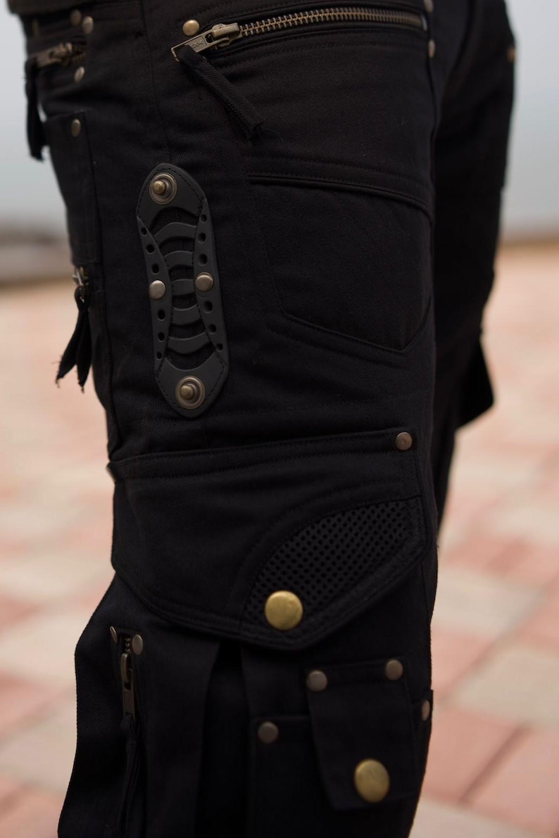 May include: Close-up of black cargo pants with multiple pockets, zippers, and decorative metal rivets. The pants feature a unique design with a mesh pocket and a leather accent piece. The overall style is utilitarian and edgy.