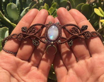 Puede incluir: Un brazalete de macramé marrón con un dije de media luna y un gran colgante ovalado de piedra lunar. El brazalete está decorado con pequeñas cuentas verdes.
