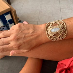 May include: A beige macrame bracelet with a white oval gemstone in the center. The bracelet is tied around a person's wrist.