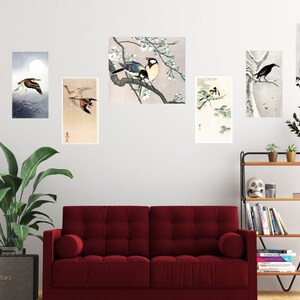 May include: A set of eight framed prints featuring Japanese-style illustrations of birds and flowers. The prints are arranged on a white wall above a red couch and a wooden shelf with books and decorative items.