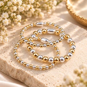 May include: A set of three stretch bracelets composed of alternating polished gold-tone and silver-tone spherical beads. Each bracelet features a single elongated, smooth metallic bead integrated into the circular design of small beads.