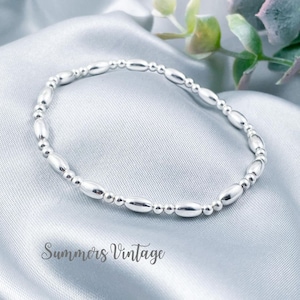 May include: A silver beaded bracelet with oval-shaped beads. The bracelet is on a light blue satin fabric background.