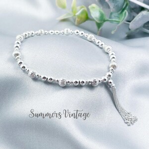 May include: A silver beaded bracelet with a tassel. The bracelet has small, round beads and larger, textured beads. A tassel hangs from the end. The text "Summers Vintage" is visible on the surface.