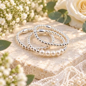May include: A set of three stretch bracelets featuring polished silver-tone metallic beads and a single focal point of five smooth white pearls on one bracelet. The beads are uniform in size, creating a reflective and classic circular design.