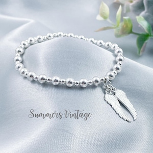 May include: A silver beaded bracelet with a silver angel wing charm. The bracelet is on a light blue satin background.