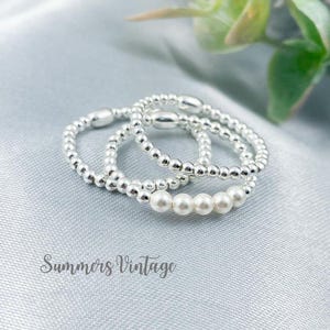 May include: Three silver rings are shown on a light blue surface. Two rings are made of small silver beads, and the third features a row of white pearls. The text "Summers Vintage" is at the bottom.