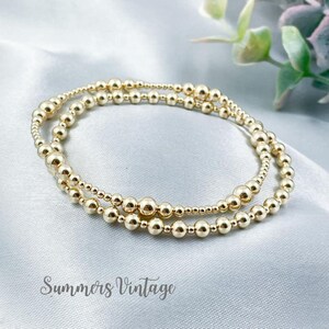 May include: Two gold beaded bracelets with different sized beads. The bracelets are layered on top of each other.