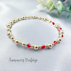 May include: A gold beaded bracelet with red crystal accents. The bracelet is made of small, round gold beads and features a few red crystal beads spaced out along the bracelet.