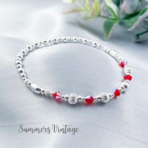 May include: A silver bracelet with small silver beads and red crystal accents. The bracelet is on a white fabric background.