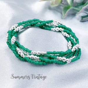 May include: A multi-strand bracelet featuring small, round, vibrant green beads interspersed with polished, reflective silver-toned metallic beads. The circular accessory is arranged in a layered, coiled design, showcasing a consistent pattern of green and silver elements throughout the entire piece.