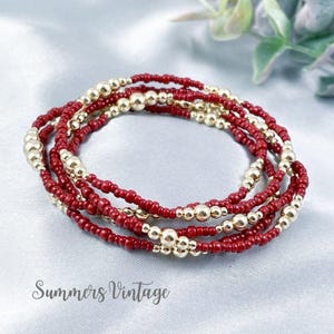 May include: A stack of three bracelets featuring small, round, deep red beads interspersed with polished gold-tone metallic beads. The bracelets are arranged in a circular formation, showcasing a consistent pattern of red and gold spheres throughout.
