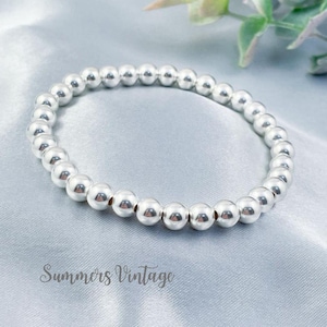 May include: A silver beaded bracelet with an elasticated design. The bracelet features small, round, shiny silver beads. It is displayed on a light blue satin fabric. The text "Summers Vintage" is visible at the bottom of the image.
