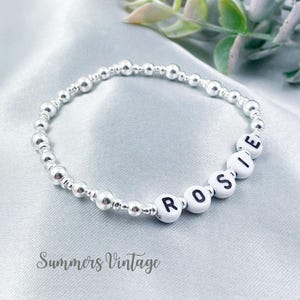 May include: A silver beaded bracelet spelling "ROSIE" with white letter beads. The bracelet is displayed on a light blue satin surface. The words "Summers Vintage" are printed in cursive at the bottom.