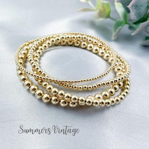 May include: A set of four gold beaded bracelets stacked on top of each other. The bracelets are made of small, round beads and are a popular style of jewellery.