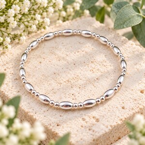 May include: A circular bracelet composed of alternating polished silver-toned beads, featuring a repeating pattern of elongated oval-shaped beads separated by smaller spherical beads, creating a continuous metallic loop with a reflective surface.